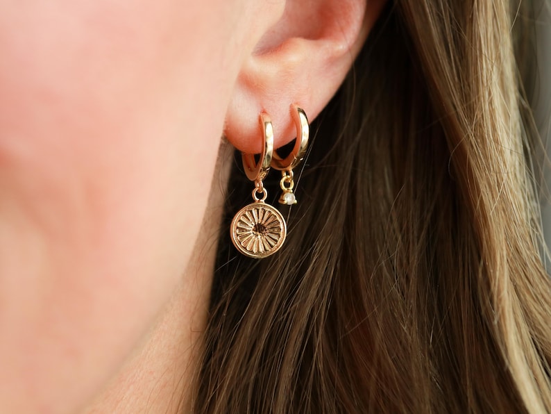 May include: Gold hoop earrings with a small gold charm hanging from one of the hoops. The charm is a round, detailed design with a small, clear stone in the center.