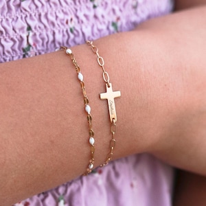 May include: Two gold-toned bracelets. One features a delicate chain with small white enamel accents. The other is a link chain with a horizontal cross charm engraved with the name Sophia. Both bracelets are worn together on a wrist.