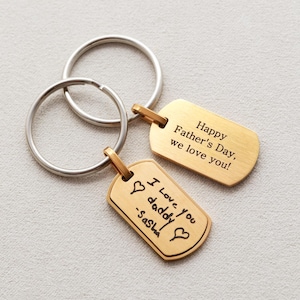 May include: Two gold-toned metal dog tag keychains with silver-toned split rings. One tag is engraved with "Happy Father's Day, we love you!" and the other features a handwritten-style engraving that reads "I love you daddy -Sasha" with two hearts.