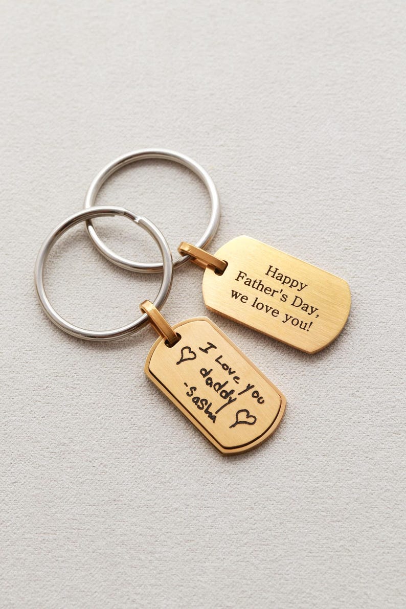 May include: Two gold-toned metal dog tag keychains with silver-toned split rings. One tag is engraved with "Happy Father's Day, we love you!" and the other features a handwritten-style engraving that reads "I love you daddy -Sasha" with two hearts.