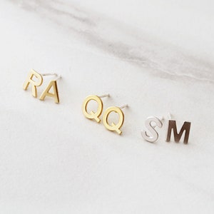May include: Three pairs of gold and silver initial earrings. The first pair is the letters "RA", the second pair is the letters "QQ", and the third pair is the letters "SM".
