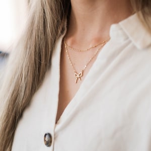 May include: A woman wearing a white button-down shirt with two gold necklaces. The top necklace has a small bow pendant.