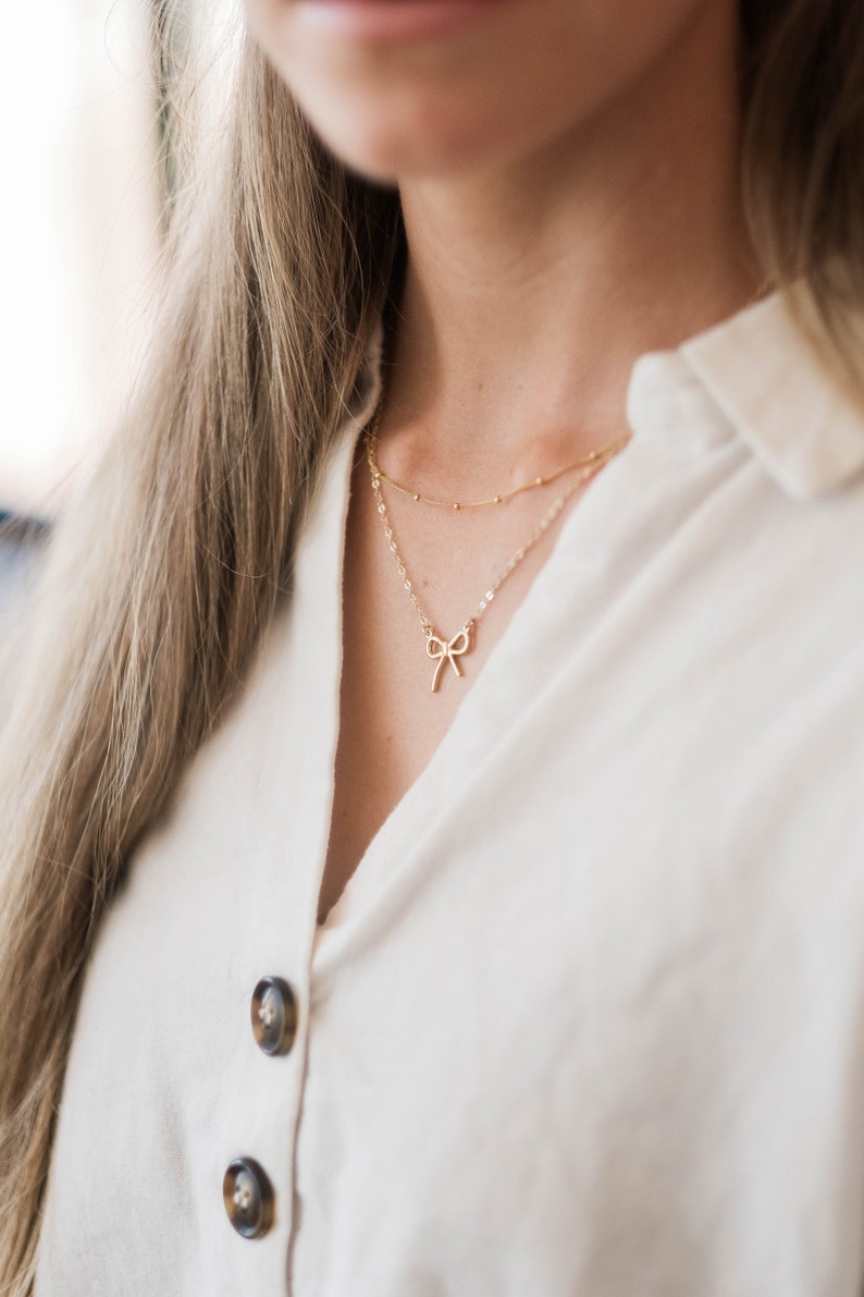May include: A woman wearing a white button-down shirt with two gold necklaces. The top necklace has a small bow pendant.