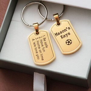 May include: Two gold-toned metal dog tag keychains with silver-colored split rings. One tag is engraved with 'Mason's Keys' and a soccer ball icon. The other tag features the text: '1. Be safe, 2. Have fun, 3. Home on time, 4. We love you'.