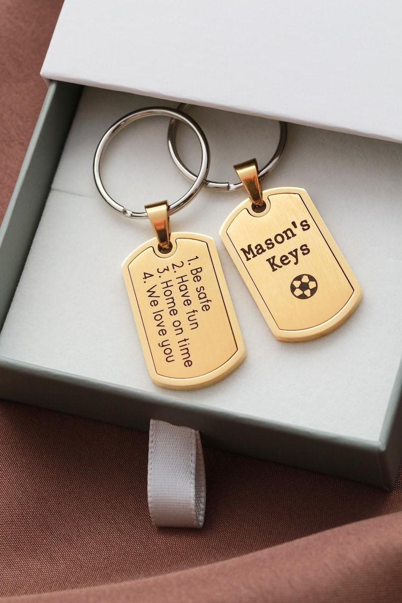 May include: Two gold-toned metal dog tag keychains with silver-colored split rings. One tag is engraved with 'Mason's Keys' and a soccer ball icon. The other tag features the text: '1. Be safe, 2. Have fun, 3. Home on time, 4. We love you'.