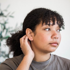 May include: A person with curly brown hair wearing a gray t-shirt and a gold chain necklace. They are wearing a gold hoop earring in their left ear and a gold bracelet on their left wrist.