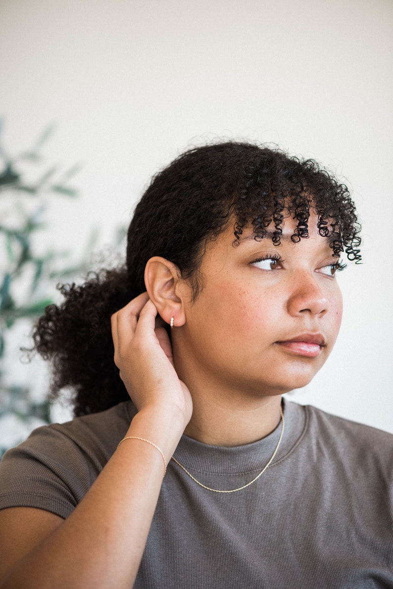 May include: A person with curly brown hair wearing a gray t-shirt and a gold chain necklace. They are wearing a gold hoop earring in their left ear and a gold bracelet on their left wrist.