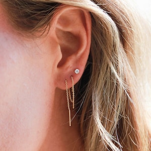 May include: Close-up of an ear with three gold earrings. A small, round, diamond stud earring is in the upper ear. Below, two gold chain earrings hang down, each with a vertical bar at the end. Blonde hair frames the ear.