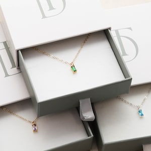 May include: Three open boxes with delicate gold necklaces featuring small, colorful gemstone pendants. The necklaces are displayed in individual boxes, each with a subtle gray background and a single letter 'D' printed on the top.