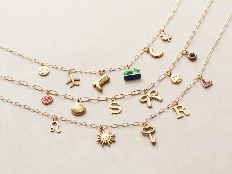 May include: Three gold-colored charm necklaces with various pendants, including a paw print, airplane, cowboy boot, strawberry, seashell, letter "S", bow, letter "R", sun, and key. The charms are attached to a delicate chain.