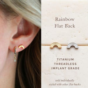 May include: Gold and silver rainbow-shaped stud earrings. The earrings are displayed next to the ear of a person wearing a gold hoop earring with a small charm. The text on the right says "Rainbow Flat Back" and "Titanium Threadless Implant Grade."