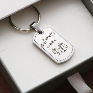 May include: A silver-toned metal dog tag keychain featuring a brushed finish. The surface is engraved with the text "Adilano BUBBE" alongside a small heart symbol and a simple line drawing of two figures standing side by side with long hair.