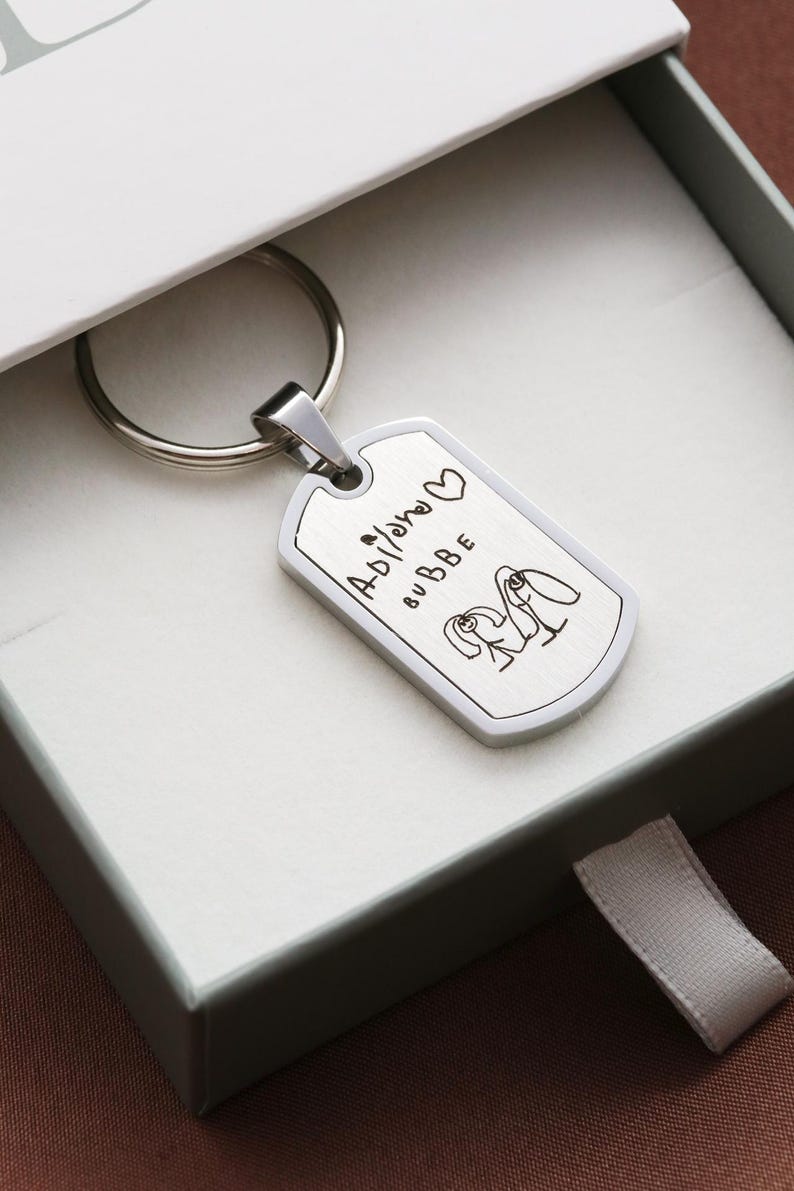 May include: A silver-toned metal dog tag keychain featuring a brushed finish. The surface is engraved with the text "Adilano BUBBE" alongside a small heart symbol and a simple line drawing of two figures standing side by side with long hair.