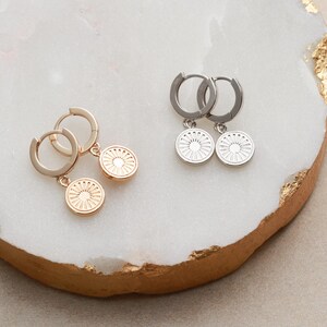 May include: Two pairs of hoop earrings with a circular pendant. One pair is gold with a gold pendant and the other pair is silver with a silver pendant. The pendants have a sunburst design.