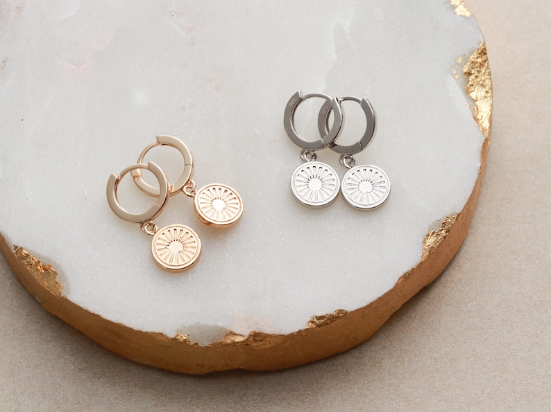 May include: Two pairs of hoop earrings with a circular pendant. One pair is gold with a gold pendant and the other pair is silver with a silver pendant. The pendants have a sunburst design.