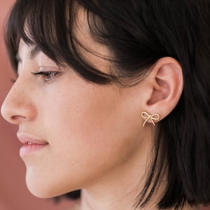 May include: A woman with short black hair wearing gold bow earrings.