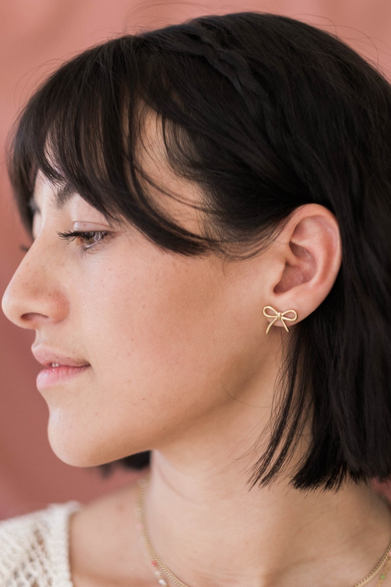 May include: A woman with short black hair wearing gold bow earrings.