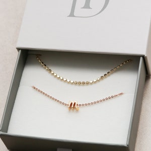 May include: A gold chain necklace with a small gold letter 'M' charm, inside a white box with the text 'TD' on the top.