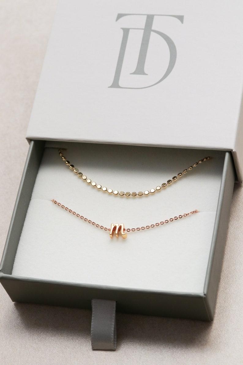 May include: A gold chain necklace with a small gold letter 'M' charm, inside a white box with the text 'TD' on the top.