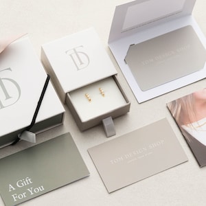 May include: A white gift box with a drawer containing a pair of gold earrings. The box has the initials "TD" embossed on the top. The gift box is surrounded by other gift packaging items, including a card that says "A Gift For You" and a card that says "TOM DESIGN SHOP".