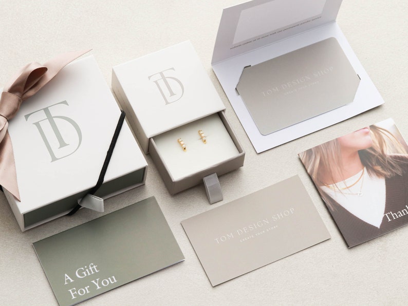May include: A white gift box with a drawer containing a pair of gold earrings. The box has the initials "TD" embossed on the top. The gift box is surrounded by other gift packaging items, including a card that says "A Gift For You" and a card that says "TOM DESIGN SHOP".