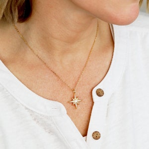May include: A gold necklace with a starburst pendant featuring a white stone in the center. The necklace is worn by a person with a white shirt and gold earrings with a starburst design.