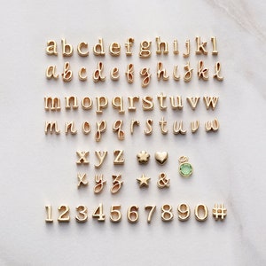 May include: Gold-toned alphabet letter charms, numbers, and symbols including a heart, star, clover, and ampersand. There is also a green gemstone charm.