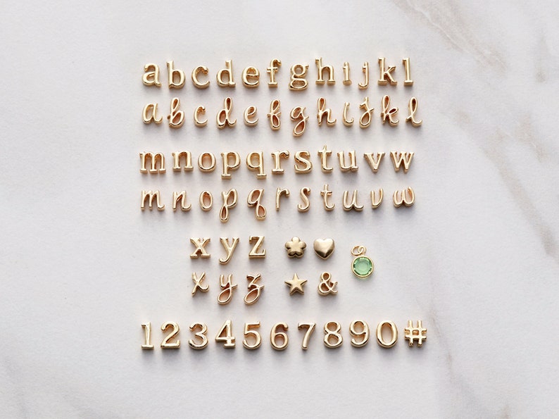 May include: Gold-toned alphabet letter charms, numbers, and symbols including a heart, star, clover, and ampersand. There is also a green gemstone charm.