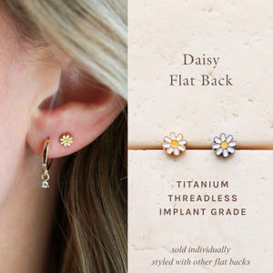 May include: Daisy flat back stud earring featuring a white and yellow flower design. The text reads Daisy Flat Back, Titanium Threadless Implant Grade, sold individually, styled with other flat backs. The earring is shown worn in an earlobe.