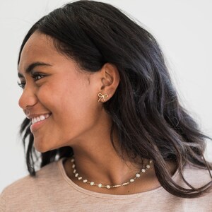 May include: A woman with long dark hair is wearing a light brown shirt and a delicate gold necklace with small white flower accents. She is also wearing a pair of small gold bow earrings.