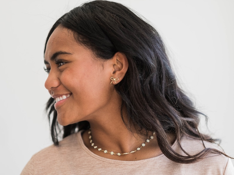 May include: A woman with long dark hair is wearing a light brown shirt and a delicate gold necklace with small white flower accents. She is also wearing a pair of small gold bow earrings.