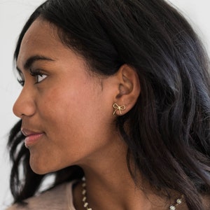 May include: A woman with long dark hair wearing a gold bow earring and a gold daisy chain necklace.
