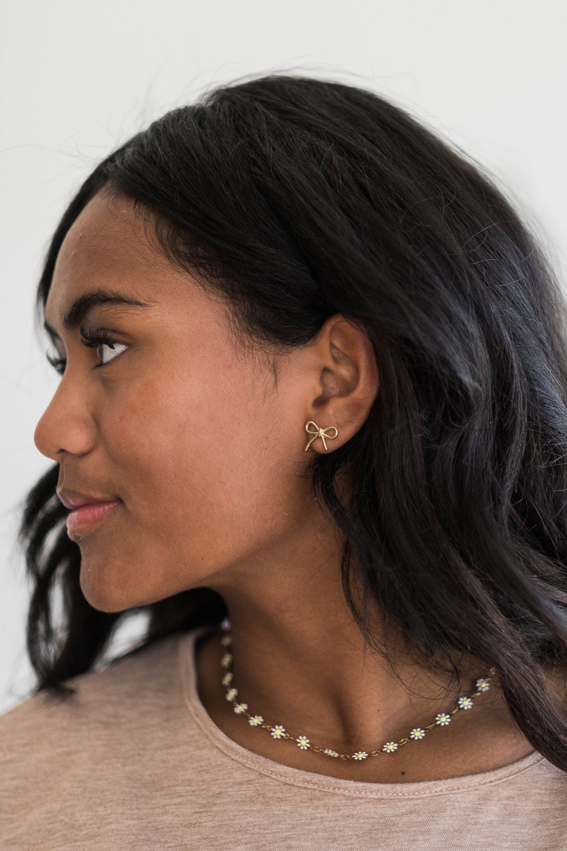 May include: A woman with long dark hair wearing a gold bow earring and a gold daisy chain necklace.
