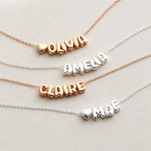May include: Four silver and gold necklaces with heart charms and names spelled out in bubble letters. The names are Olivia, Amelia, Claire, and Mae.