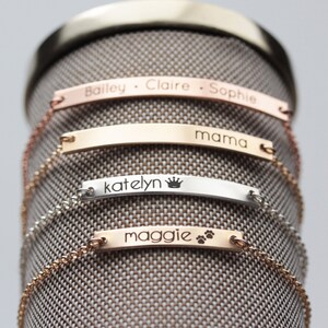 May include: Four personalized silver and gold bar bracelets with chains. The bracelets are engraved with the names "Bailey, Claire, Sophie", "mama", "katelyn" with a crown symbol, and "maggie" with paw prints.