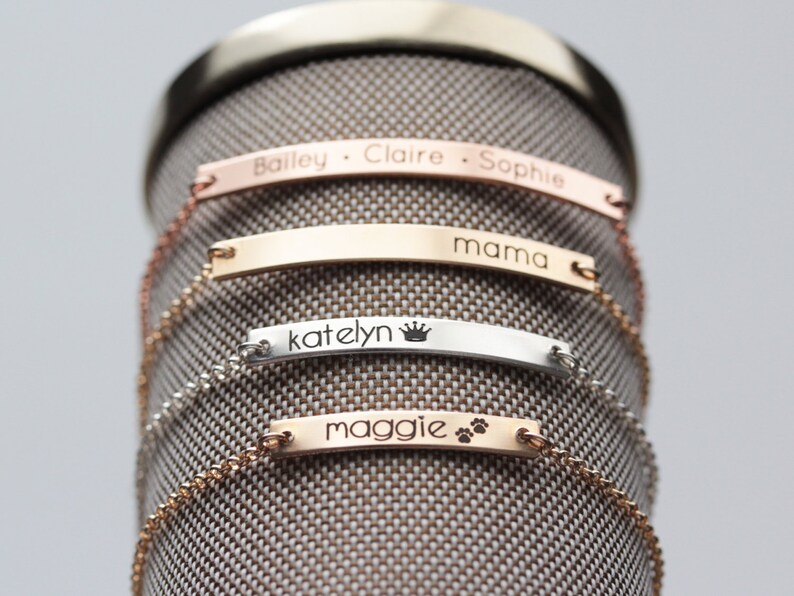 May include: Four personalized silver and gold bar bracelets with chains. The bracelets are engraved with the names "Bailey, Claire, Sophie", "mama", "katelyn" with a crown symbol, and "maggie" with paw prints.