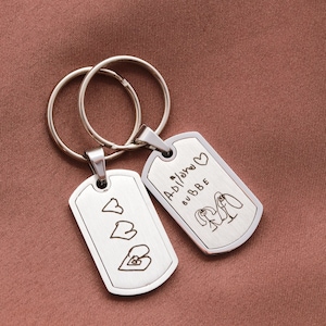 May include: Two silver-toned metal dog tag keychains with split rings. One tag features three engraved heart outlines. The other tag displays the engraved text "Adilano BUBBE" with a heart symbol and a line drawing of two figures holding hands.