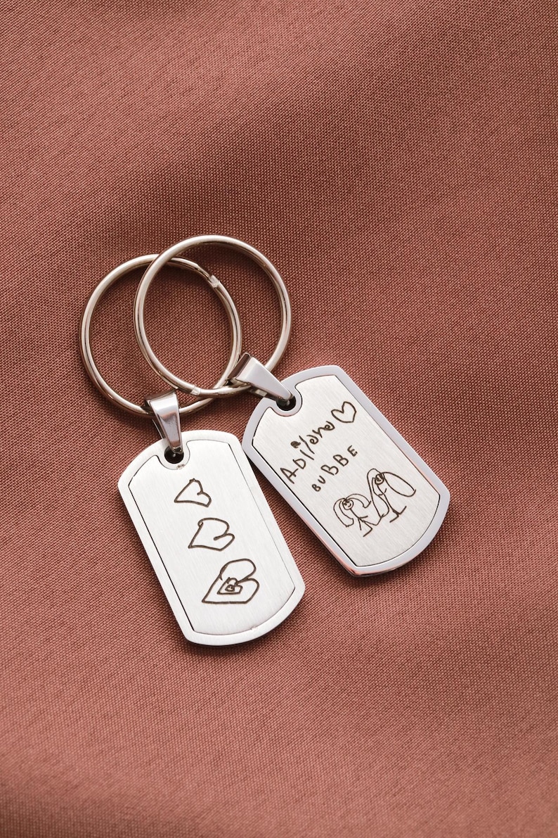May include: Two silver-toned metal dog tag keychains with split rings. One tag features three engraved heart outlines. The other tag displays the engraved text "Adilano BUBBE" with a heart symbol and a line drawing of two figures holding hands.