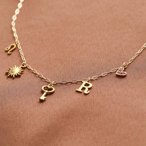 May include: A gold-colored necklace with multiple charms. The charms include a sun, a key, the letter "R", a heart with pink stones, and a Leo zodiac symbol. The necklace is displayed on a brown surface.