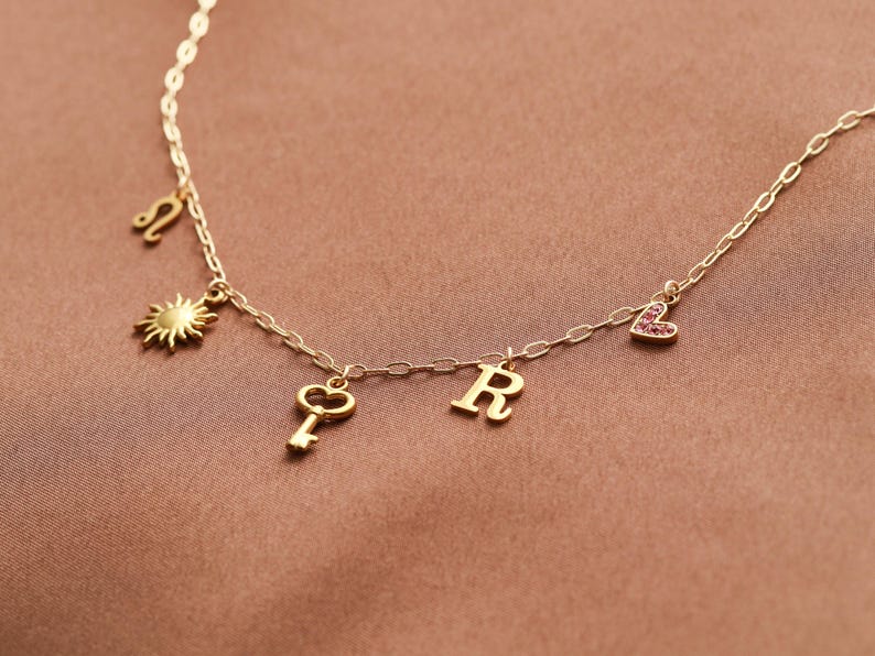 May include: A gold-colored necklace with multiple charms. The charms include a sun, a key, the letter "R", a heart with pink stones, and a Leo zodiac symbol. The necklace is displayed on a brown surface.