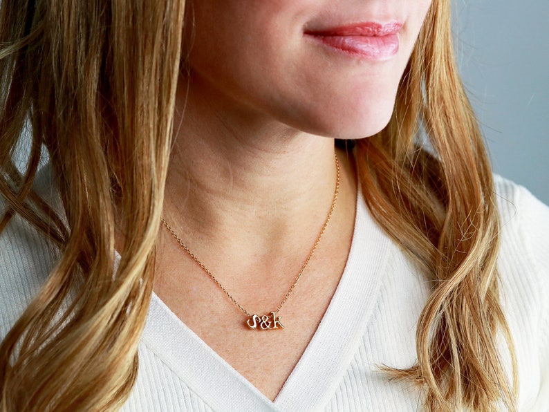 May include: A gold necklace with a delicate chain and a pendant featuring the letters "S&K" in a cursive font.