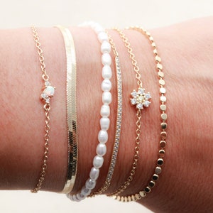 May include: A set of five gold bracelets with different chain styles. One bracelet features a small opal stone with diamond accents, another has a flower-shaped charm with diamonds, and another has a row of small pearls. The remaining two bracelets are simple gold chains.