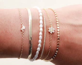 May include: A set of five gold bracelets with different chain styles. One bracelet features a small opal stone with diamond accents, another has a flower-shaped charm with diamonds, and another has a row of small pearls. The remaining two bracelets are simple gold chains.