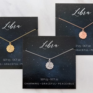 May include: Three necklaces with zodiac signs on them. The necklaces are on black cards with the zodiac sign Libra, the dates September 23 - October 22, and the words "Charming, Graceful, Peaceable". The necklaces are gold, rose gold, and silver.