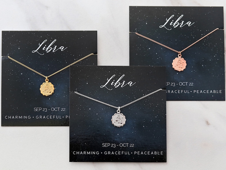 May include: Three necklaces with zodiac signs on them. The necklaces are on black cards with the zodiac sign Libra, the dates September 23 - October 22, and the words "Charming, Graceful, Peaceable". The necklaces are gold, rose gold, and silver.