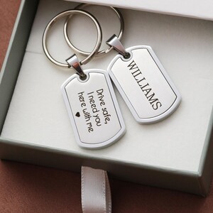 May include: Two silver-toned metal dog tag keychains with split rings. One tag is engraved with the text "WILLIAMS" and the other is engraved with "Drive safe, I need you here with me" followed by a small heart symbol. The keychains are inside a box.