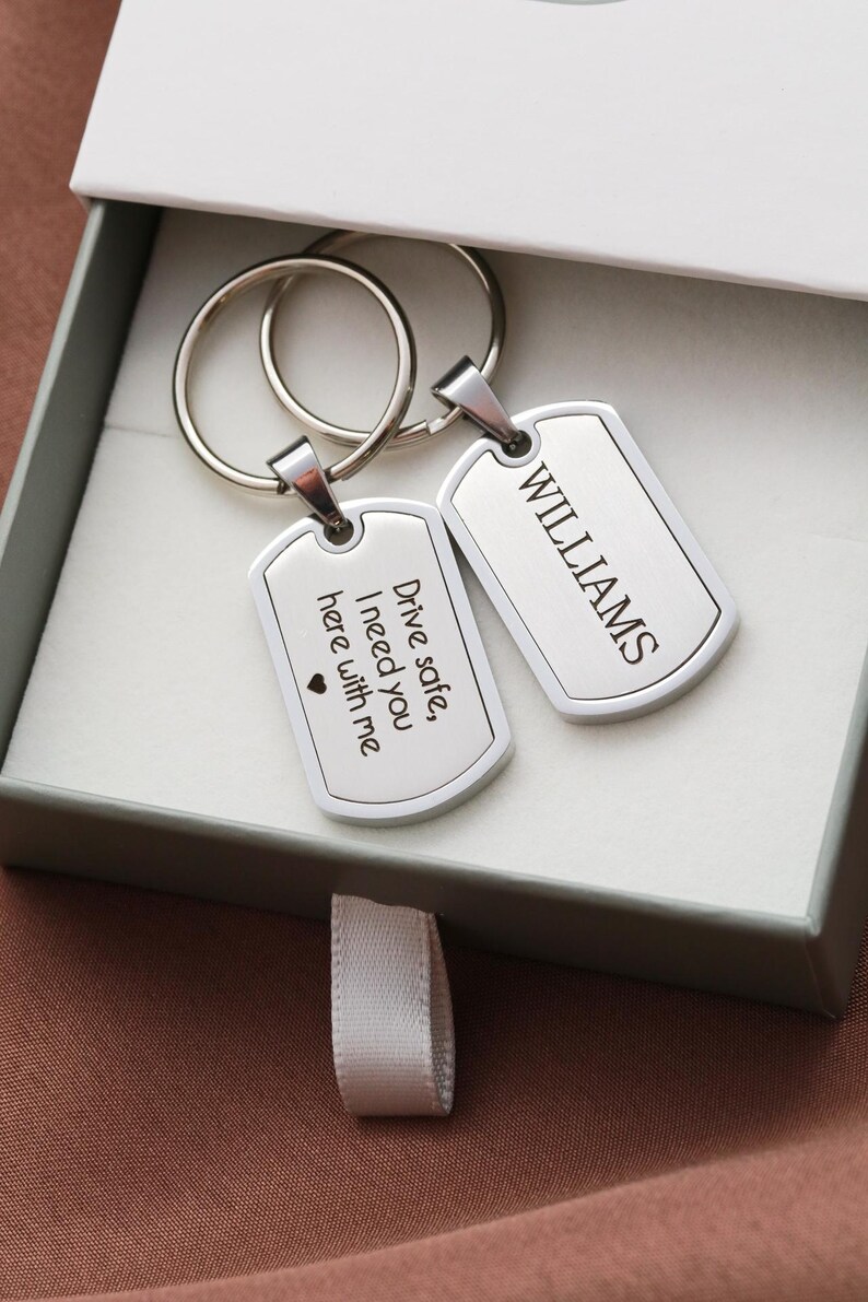May include: Two silver-toned metal dog tag keychains with split rings. One tag is engraved with the text "WILLIAMS" and the other is engraved with "Drive safe, I need you here with me" followed by a small heart symbol. The keychains are inside a box.