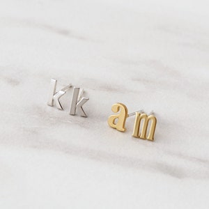 May include: Two pairs of initial earrings. One pair is silver and features the letter "K" twice. The other pair is gold and features the letters "a" and "m".
