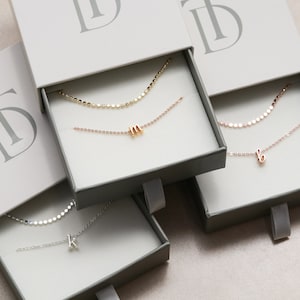 May include: Three open boxes with gray interiors and white lids. Each box contains a delicate silver, gold, or rose gold chain necklace with a small letter charm. The boxes have a logo on the lid that says "TD".