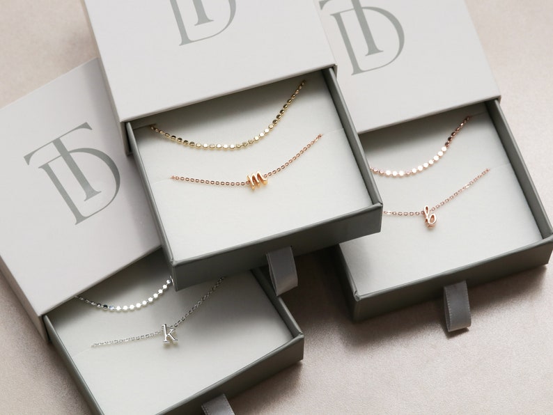 May include: Three open boxes with gray interiors and white lids. Each box contains a delicate silver, gold, or rose gold chain necklace with a small letter charm. The boxes have a logo on the lid that says "TD".